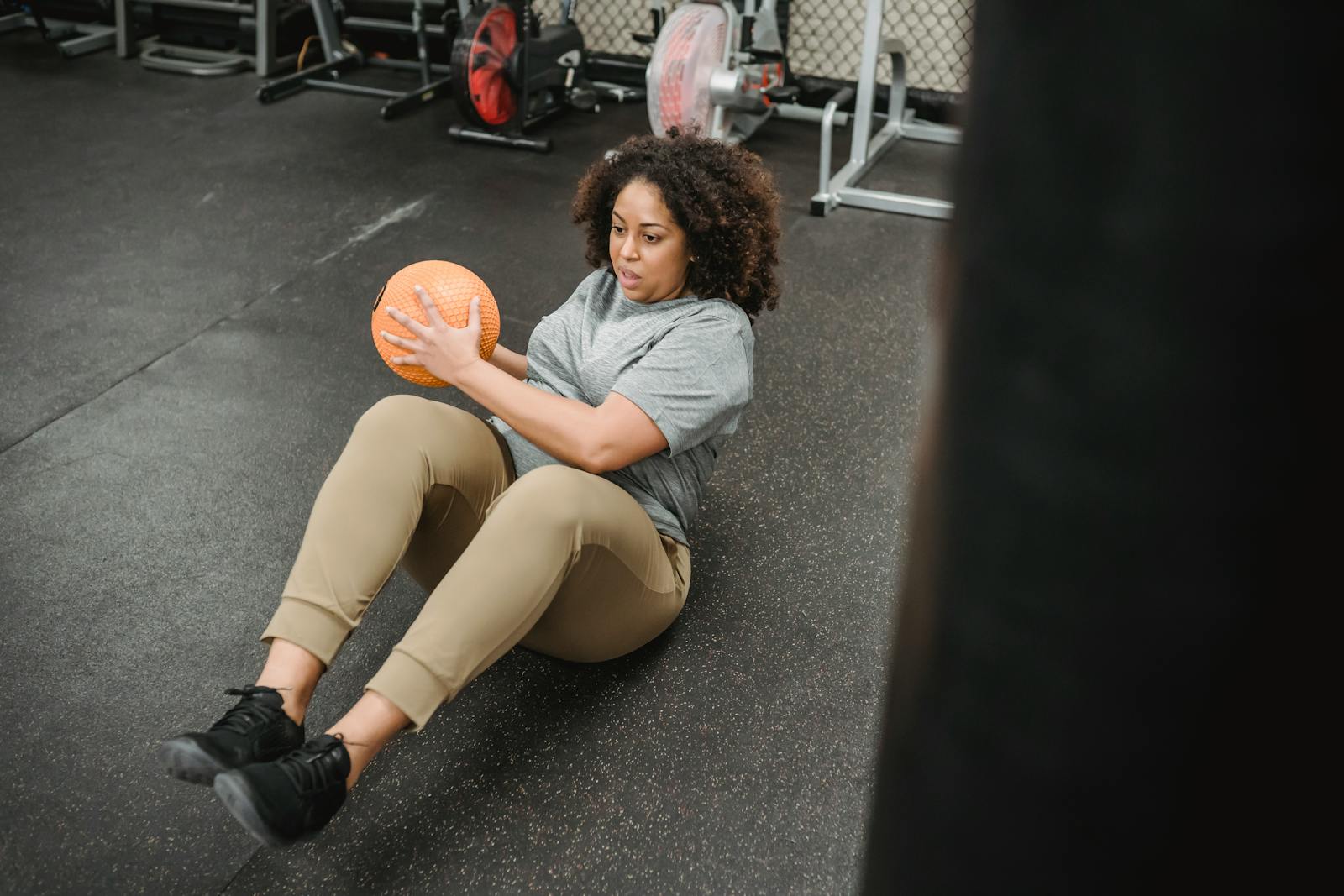 A woman performs a seated twist with a medicine ball in an indoor gym setting, demonstrating strength and focus.